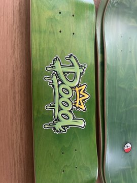 Close-up of green skateboard decks leaning against a wooden surface, one featuring a stylized 'HOOD' graffiti-style logo with a yellow star.