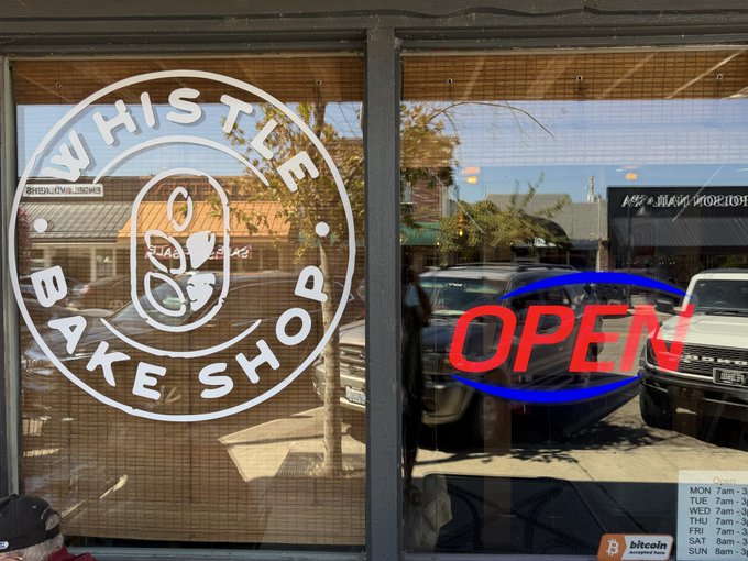 The image shows the storefront window of "Whistle Bake Shop" with an "OPEN" sign and a Bitcoin accepted here sticker visible in the corner. This relates to the post by showcasing the local coffee shop where the customer purchased coffee and baked goods while paying with Bitcoin, with their shop hours (Mon-Sat 7am-3pm) displayed on the door.