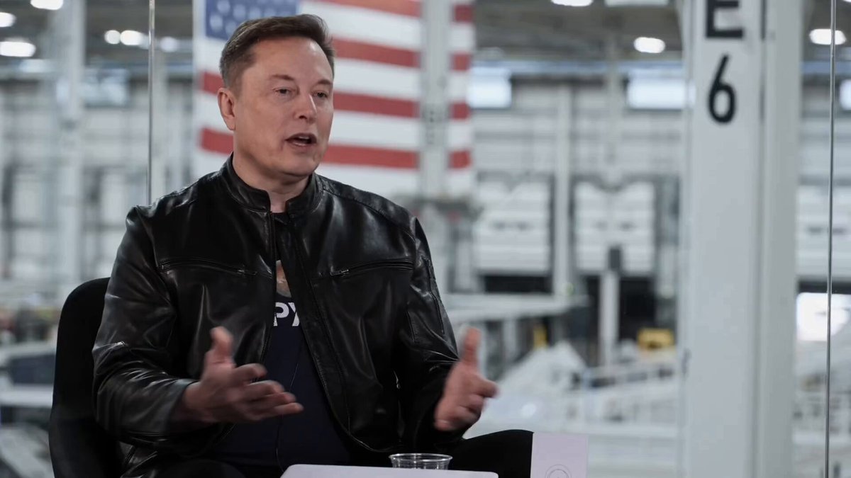 The image shows an interview-style photo featuring a man wearing a black leather jacket, sitting at a desk with his hands outstretched as if he's explaining something or making a point. The background of the image reveals that they are inside a building, possibly a factory or warehouse, and there is a large American flag visible in the scene. The caption below the photo reads "Elon Musk explains his timeline".