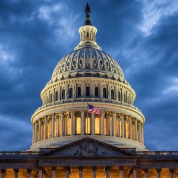 The illuminated dome of the US Capitol building stands against a deep blue twilight sky, with the American flag visible below.