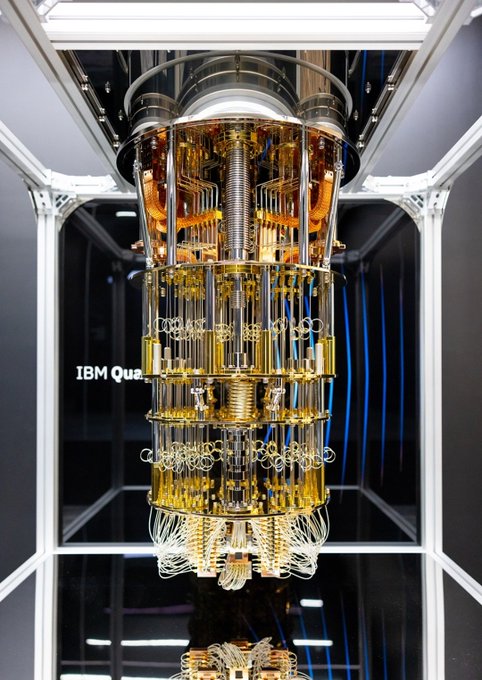 A close-up view of an IBM Quantum computer's chandelier-like cryogenic cooling structure, featuring gold-colored wiring and components, with 'IBM Qua' visible on the side.