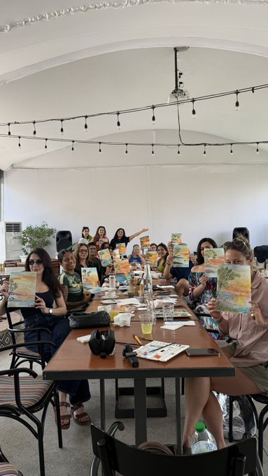 A group of people sitting around a long wooden table in a white tent venue, holding up their finished landscape paintings during what appears to be a paint and sip class.