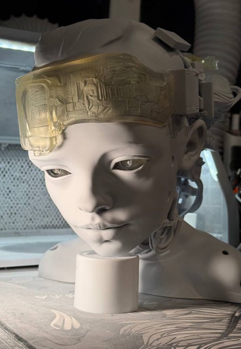 A close-up photo of a robotic mannequin head with detailed facial features, wearing a yellow translucent headband device, with mechanical components visible behind the head.