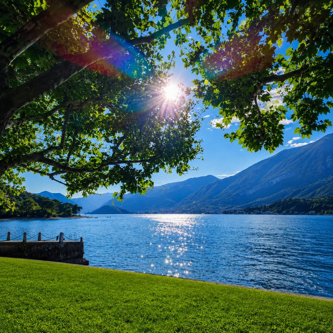 The image shows a beautiful lake surrounded by mountains under a clear blue sky with sunlight shining through the trees. The sun's rays are reflecting off the water, creating a stunning visual effect. In the foreground of the photo, there is a grassy area near the shoreline where people can sit and enjoy the view or engage in outdoor activities like walking along the lake or having a picnic by the water.