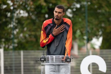 Victor Wembanyama stands at a podium wearing an orange and black Spurs tracksuit, with his hand placed over his heart as he speaks at the inauguration of the newly renovated basketball court in Nanterre, his hometown.