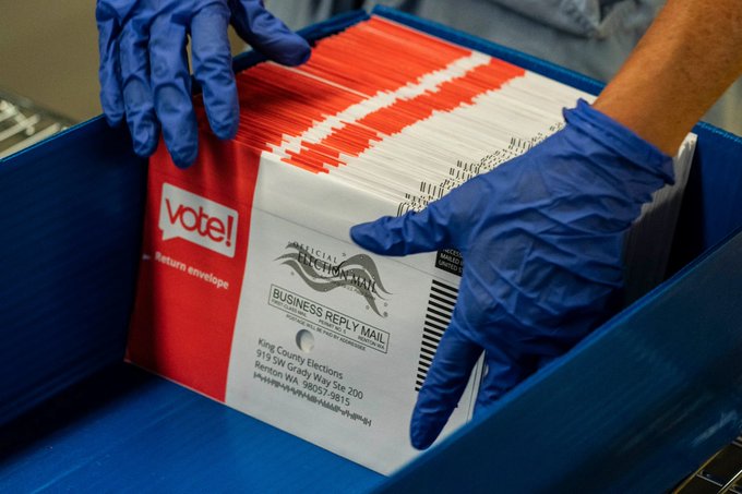 Gloved hands handling a stack of vote return envelopes in a blue bin, with the visible envelope showing 'vote!', 'King County Elections, 919 SW Grady Way Ste 300, Renton WA 98057-9815', and a 'Business Reply Mail' stamp.