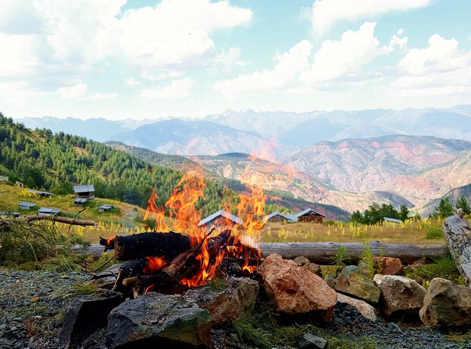A campfire burns in a fire ring in the foreground with a scenic mountain landscape and small cabins visible in the background.