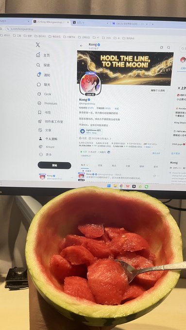 A computer monitor displays the X (Twitter) profile of 'Kong @kongzhonghong' with a banner reading 'HOLD THE LINE TO THE MOON!', while a hollowed-out watermelon bowl filled with red fruit chunks sits in front.