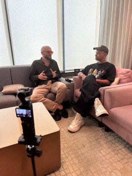 Two men sit on a pink couch in a room, having a conversation while being filmed by a camera mounted on a tripod in the foreground.