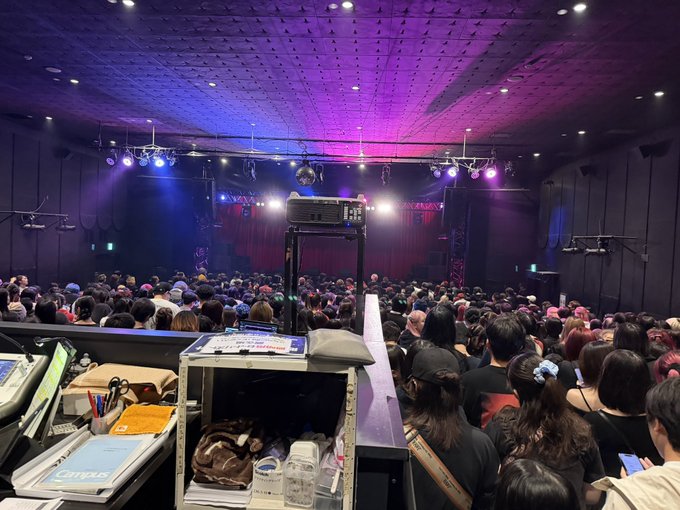 A crowded concert venue viewed from behind a sound/lighting booth, with purple stage lights illuminating the audience.