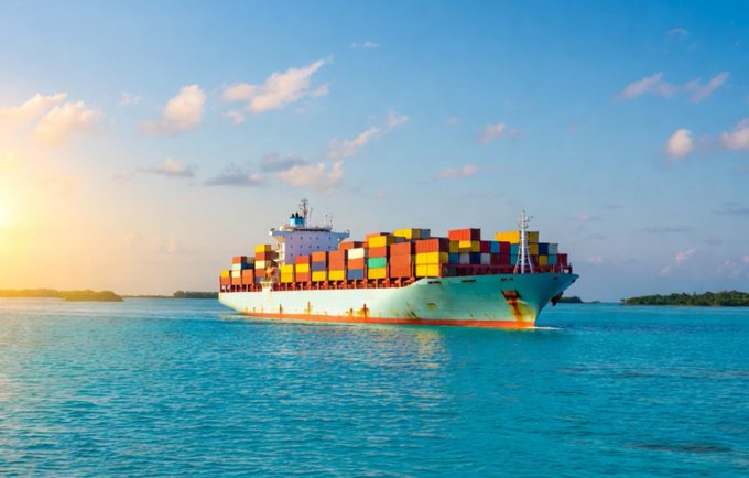 A large container ship loaded with multicolored cargo containers sails through turquoise ocean waters under a bright blue sky with scattered clouds.