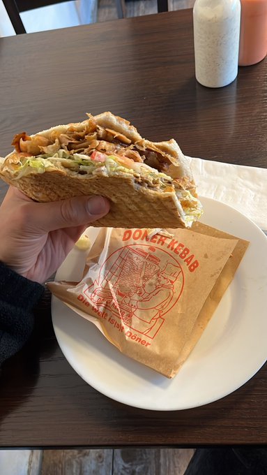 The image shows a person holding up a sandwich, which appears to be a pita or a wrap filled with meat and vegetables. The sandwich is placed on top of a brown paper bag that has red writing on it. In the background, there are two bottles and a cup, suggesting that this scene takes place in a restaurant setting.