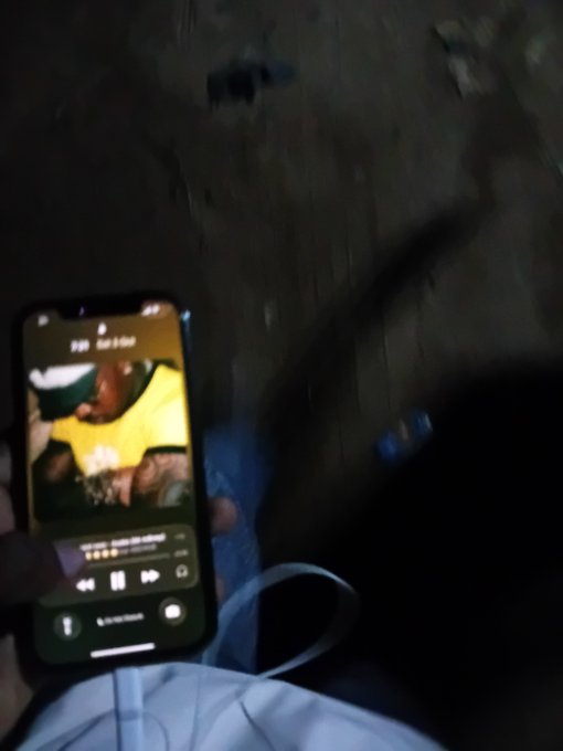 Blurry photo of a hand holding a smartphone playing music, showing an album cover with a person in a yellow shirt, with a charging cable visible.