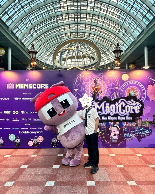 A person wearing a white cat-eared filter poses with the purple Memecore mascot in front of a 'MagiCore: A New Chapter Begins Here' event backdrop under a glass dome.