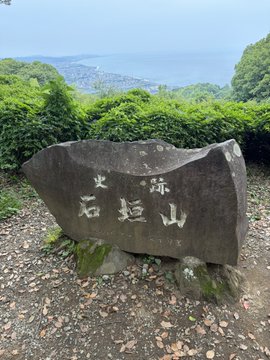 A stone monument engraved with the Japanese characters "石垣山" (Ishigakiyama) with smaller characters "史跡" (historic site) above, set against a backdrop of ocean and lush greenery.