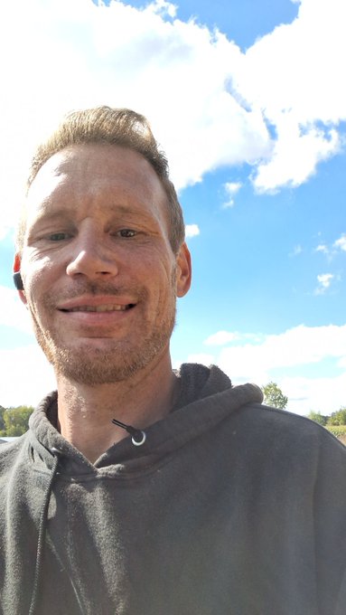 A man wearing a dark hoodie and earbuds smiles at the camera in a selfie taken outdoors under a bright blue sky.