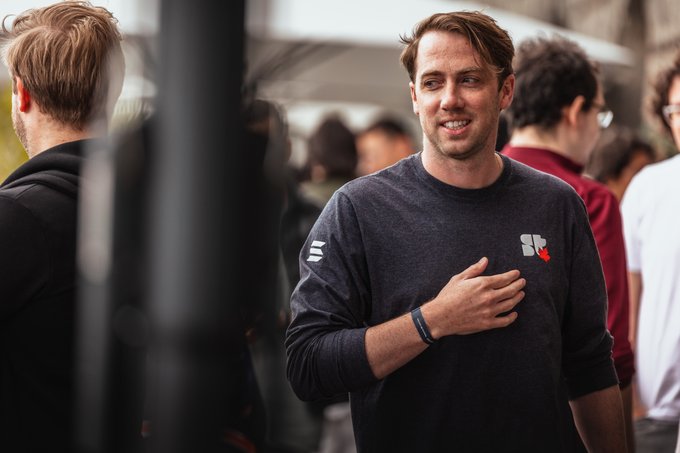 A man with reddish-brown hair wearing a dark grey long-sleeve shirt with a small red logo, smiling while standing in a crowd at an outdoor event.