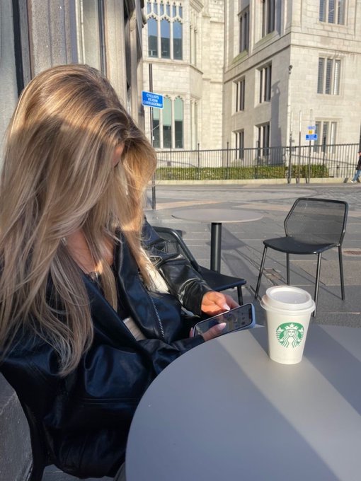 A blonde woman in a black leather jacket sits at an outdoor café table looking at her phone, with a Starbucks coffee cup beside her, in a city street setting — capturing the "gm" (good morning) vibe and on-the-go energy of starting the day in the city.