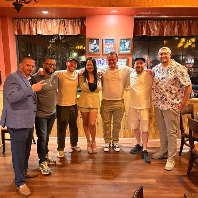A group of seven people pose together with arms around each other in a warmly lit restaurant or bar setting with peach-colored walls.