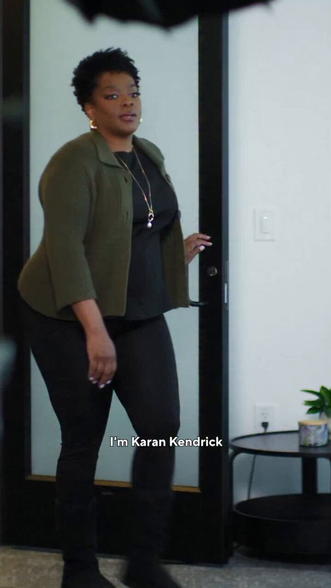 The image features a woman standing in front of a door, wearing black pants and a green jacket. She has her hand on the door handle, possibly waiting for someone or just passing by. The background shows a white wall with a plant placed next to the door. There is also an umbrella visible in the scene.