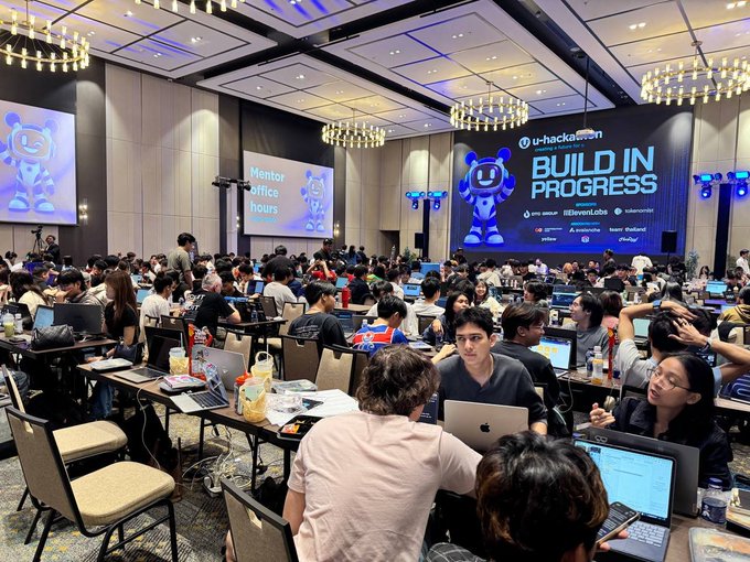 A crowded hackathon setting with many participants working on laptops; large screens display 'U-hackathon', 'BUILD IN PROGRESS', and 'Mentor office hours'.