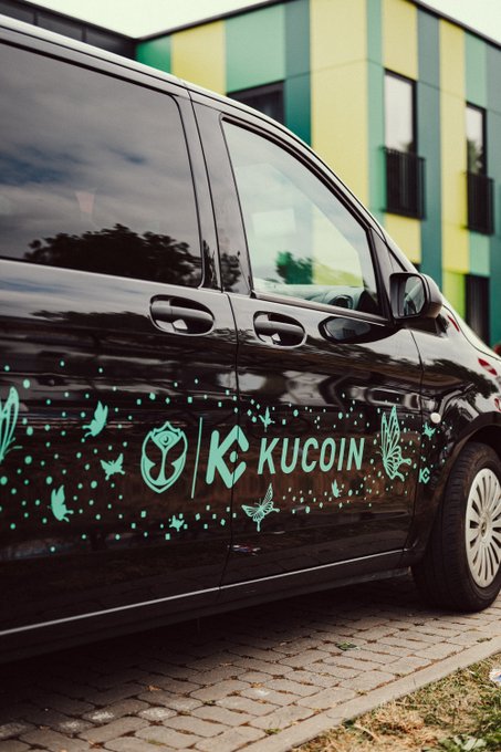 A black van displays the KuCoin cryptocurrency exchange logo alongside decorative teal butterflies and sparkles, parked outside a modern building—tying into the Tomorrowland festival theme through its butterfly motifs.