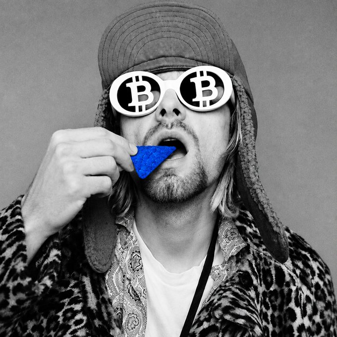 A black-and-white photo shows a man wearing a furry hat, leopard-print jacket, and sunglasses printed with the Bitcoin (₿) logo, holding a blue triangular object to his mouth.