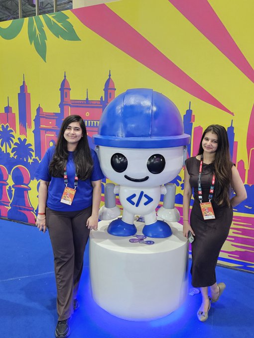 Two women pose on either side of a blue and white robot mascot featuring coding symbols (< />), set against a vibrant yellow and pink backdrop with city skyline silhouettes.