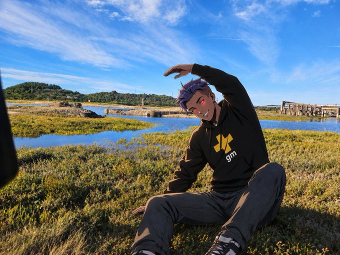 An anime-styled figure of a purple-haired character in a black hoodie with a yellow logo sits on grass in a wetland area, posing with one hand raised against a blue sky with wispy clouds.