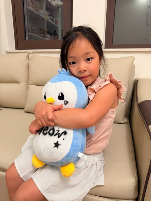 A young girl happily hugs a blue and white Pudgy Penguin plush toy (with "meow" text visible) on a beige couch, capturing the wholesome spirit of the post as the next generation embraces the beloved NFT brand in a tangible, cuddly form.
