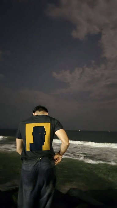 A man seen from behind stands on a beach at night, wearing a t-shirt with a pixel-art style graphic in yellow and dark blue.