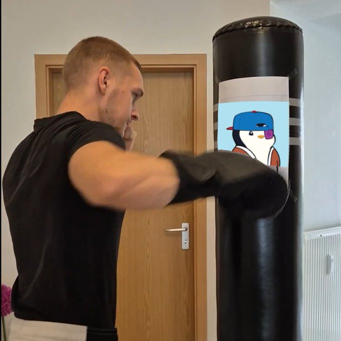 A man in a black t-shirt and boxing gloves punches a black heavy bag that features a cartoon penguin logo wearing a red and blue cap.