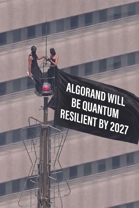 Two people on an elevated platform attached to a building, hanging a black banner that reads 'ALGORAND WILL BE QUANTUM RESILIENT BY 2027'.