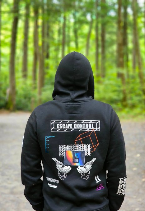 A person wearing a black hoodie with their hood up, photographed from behind in a forest setting; the hoodie's back features an 'ESCAPE CONTROL' design with illustrations of hands, a cube, and digital graphics.