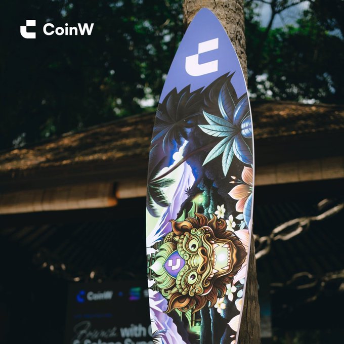 A CoinW-branded surfboard with vibrant tropical artwork leans against a tree, displaying the text "CoinW" in the upper left corner. The setting reflects the post's "Crypto, but make it an adventure" theme by placing the crypto brand in an outdoor, exotic locale.