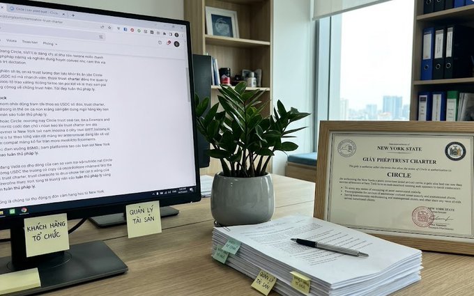 An office desk with a computer monitor displaying Vietnamese text about Piepmeist Charter, alongside sticky notes reading 'KHÁCH HÀNG TỔ CHỨC' and 'QUẢN LÝ TÀI SẢN', a potted plant, stacked documents, and a framed certificate for 'NEW YORK STATE PIEPMEIST CHARTER CIRCLE'.
