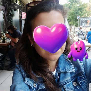 Outdoor selfie photo of a woman in a denim jacket with a large purple/pink heart emoji and a small purple cartoon monster character covering her face, taken at what appears to be an outdoor café.