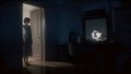 A dimly lit room where a person stands in a doorway while another sits watching a glowing Apple TV screen in the darkness.