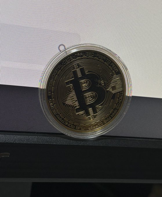 A physical Bitcoin collectible coin in its clear plastic case is displayed on a laptop, featuring the iconic "₿" symbol and inscriptions including "BITCOIN DIGITAL DECENTRALIZED PEER TO PEER," "FINE COPPER," and "TROY OUNCE." The image humorously illustrates the poster's dream of selling this novelty copper token to someone as if it were actual Bitcoin worth thousands of dollars.