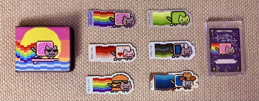 A collection of pixel-art stickers featuring Nyan Cat and its variants (like "Peel Me") displayed on a textured beige background, alongside a small boxed trading card.