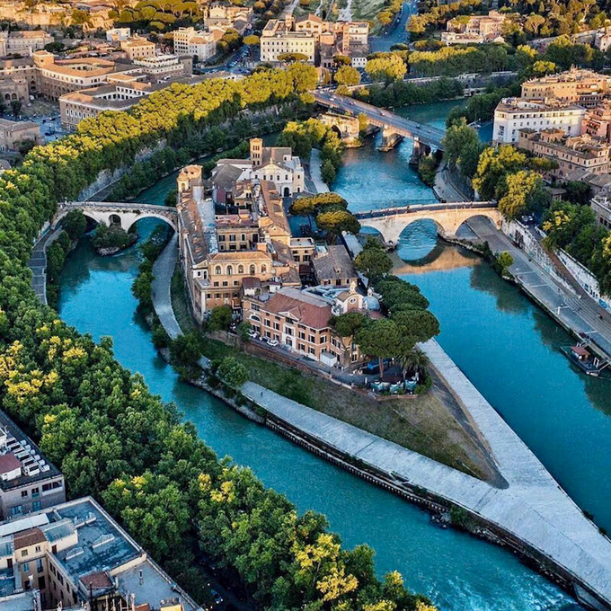 An aerial view of Tiber Island in Rome, Italy, showing historic buildings surrounded by the Tiber River with bridges connecting it to the city.