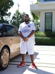 A man stands smiling next to a gold sports car, wearing a white t-shirt with "USDC" printed on it—referencing the USD Coin stablecoin. The image ties into the post's question about market sentiment by visually linking luxury and crypto, prompting viewers to weigh whether this lifestyle signals bullish optimism or a market top.
