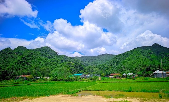 A scenic landscape view of a lush green village with rice paddies in the foreground, houses in the midground, and mountains under a cloudy blue sky.