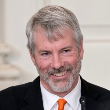 A smiling gray-haired man with a beard, wearing a dark suit and orange tie, appears to be Michael Saylor, the executive behind the Bitcoin purchasing announcement. The image likely relates to the post about Strategy (formerly MicroStrategy) $MSTR acquiring 267 additional Bitcoin.