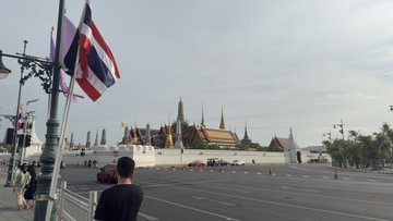 The image shows a man standing next to a street lamp near a temple in Thailand. The man appears to be looking at something or someone across the street. There are several other people visible in the scene, some of them carrying backpacks. A car is parked on the side of the road, and there's an airplane flying overhead. The image also contains text that reads "You nailed it, the one who said Thailand! We arrived in Bangkok, taxi paid with a tap on the special edition virtual Solana card right inside the airport itself".
