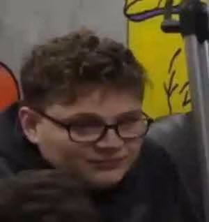 A young boy with curly hair and round glasses appears to be speaking or reacting, with a yellow background partially visible behind him.