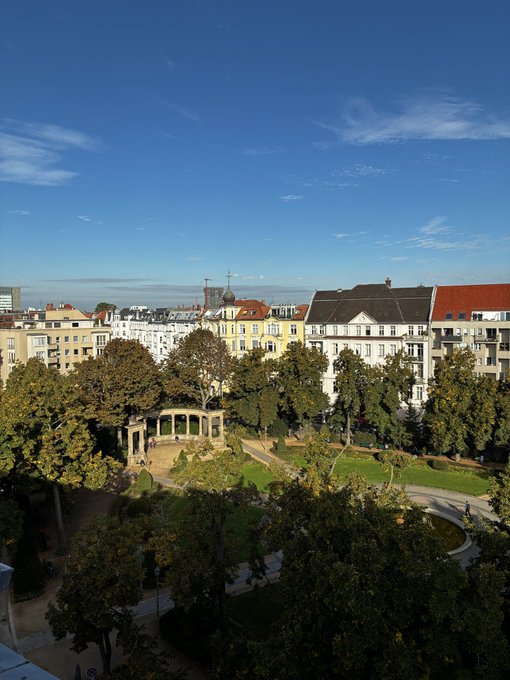 An elevated view of a European city park with green trees, a gazebo, and historic buildings with red and white facades under a clear blue sky.