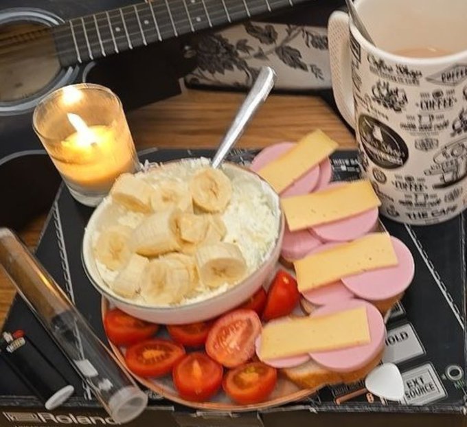 A candle-lit coffee mug sits beside a guitar on a Roland amplifier, accompanied by a plate of cheese, ham, banana-topped cream, and cherry tomatoes, symbolizing a cozy moment of creative hustle—reinforcing the message that hard work is beginning to bear fruit and inviting onlookers to join in rather than stay on the sidelines.
