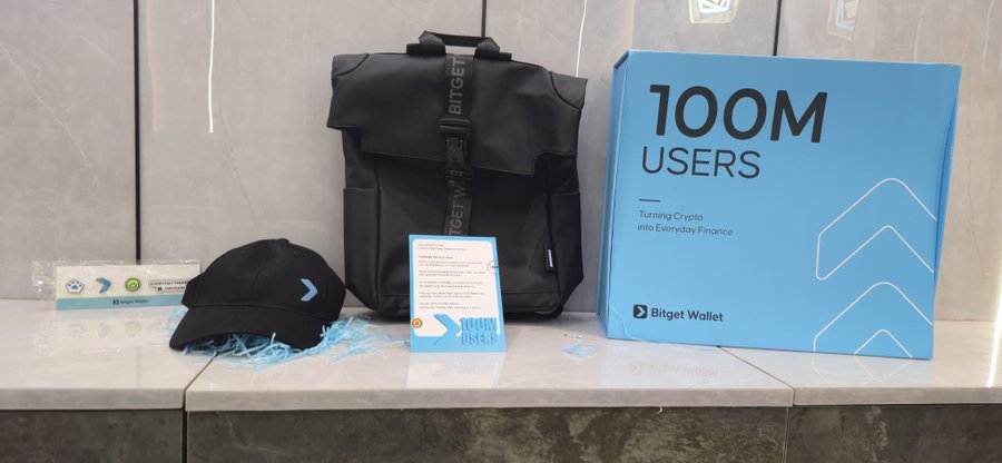 Promotional merchandise for Bitget Wallet on a counter, including a black backpack, a cap, a small card, and a blue box reading '100M USERS Turning Crypto into Everyday Finance'.