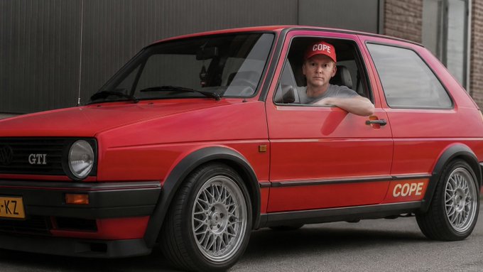 A young man wearing a red 'COPE' cap sits in a red Volkswagen GTI Mk2 with the word 'COPE' on its side, smiling at the camera.